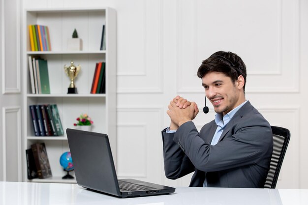 Chico lindo de servicio al cliente en traje gris con computadora portátil y auriculares tomados de la mano y sonriendo