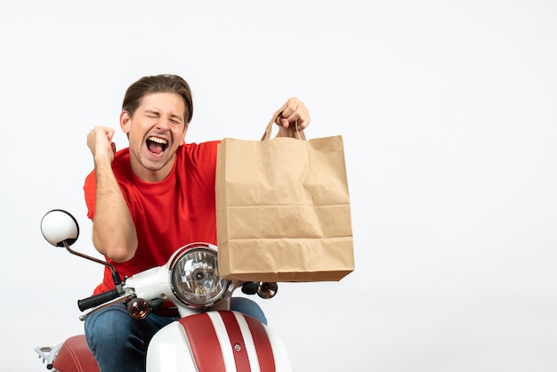 Chico mensajero sonriente joven en uniforme rojo sentado en scooter sosteniendo una bolsa de papel disfrutando de su éxito en la pared blanca