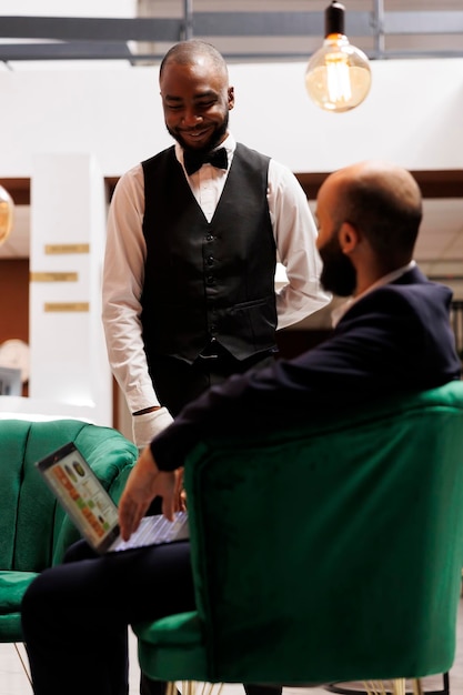 Foto gratuita chico de mesa hablando con el cliente en el salón, ayudando con los servicios de lujo mientras él espera para hacer el procedimiento de registro. hombre de negocios usando portátil para asociación de proyecto oficial, viaje de negocios.