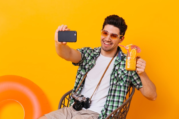 Chico morena con camisa verde toma selfie y tiene cóctel de naranja. Retrato de hombre con cámara retro posando en espacio aislado con círculo inflable.