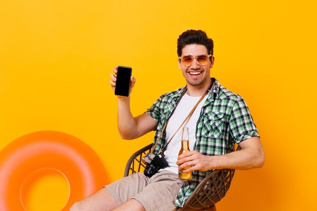 Chico morena muestra su teléfono inteligente con una sonrisa. El hombre en camisa, camiseta y pantalones cortos está sentado en una silla con una botella de cerveza en un espacio naranja con un círculo inflable.