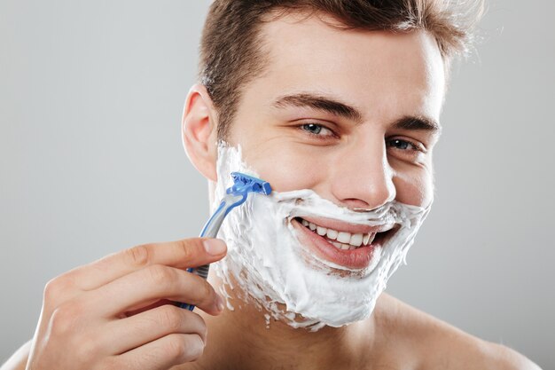 Chico moreno masculino con el pelo corto y oscuro afeitándose la cara con una maquinilla de afeitar y gel o crema satisfecha sobre la pared gris de cerca