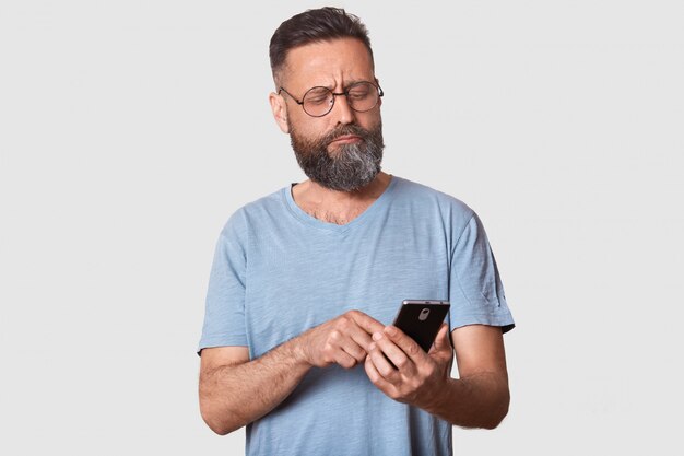 Un chico moreno tranquilo y serio mira atentamente a un lado, sosteniendo un teléfono inteligente en la mano, escribiendo mensajes a sus amigos, confundido acerca de las publicaciones. Modelo guapo con camiseta casual y especificaciones de moda.