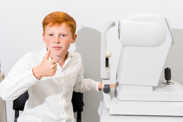Chico mostrando pulgar arriba gesto sentado cerca de la máquina de refractómetro en la clínica de óptica