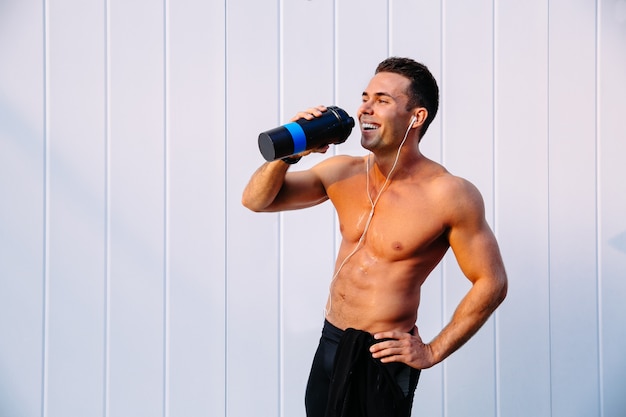 Chico musculoso alegre disfrutando de beber agua después del entrenamiento, escuchando música en auriculares