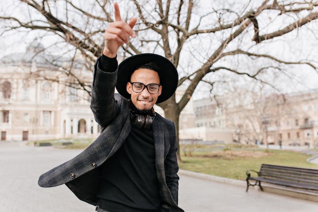Chico negro inspirado en chaqueta gris agitando la mano en el parque. Retrato al aire libre del modelo masculino africano alegre con sombrero y gafas descansando en la plaza de la ciudad.