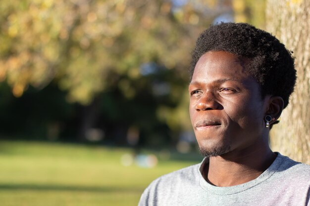 Chico negro pensativo pasando tiempo al aire libre en el parque de la ciudad. Retrato de un apuesto afroamericano con una camiseta gris mirando a un lado y pensando. Fotografía de cerca. Juventud, estilo de vida, concepto de tiempo libre.