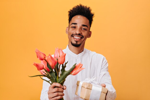 Chico de ojos marrones en camisa blanca con tulipanes rosas y regalo en pared naranja