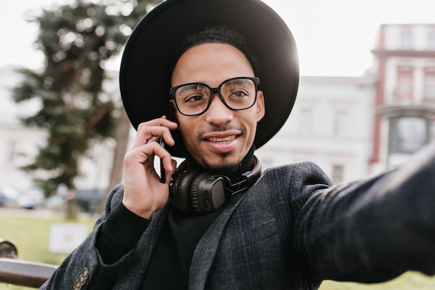 Chico de ojos oscuros en auriculares haciendo selfie en el parque. Hombre africano elegante que llama a alguien mientras toma una foto de sí mismo.