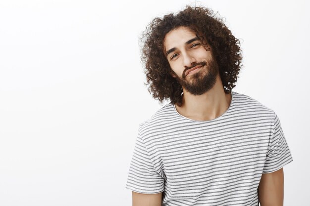 Chico oriental confiado guapo despreocupado con barba y peinado afro, inclinando la cabeza hacia un lado y sonriendo ampliamente