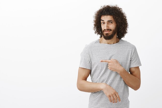 Chico oriental guapo e impresionado dudoso con barba y peinado rizado en camiseta a rayas, apuntando hacia la izquierda y levantando la ceja con expresión sospechosa e incrédula