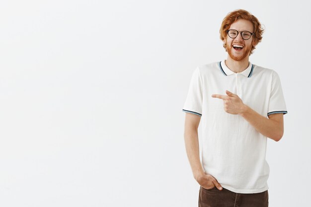 Chico pelirrojo barbudo sonriente posando contra la pared blanca con gafas