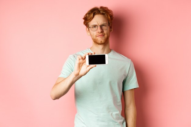 Chico pelirrojo escéptico con gafas mostrando la pantalla del teléfono inteligente horizontalmente sonriendo y frunciendo el ceño decepcionando ...