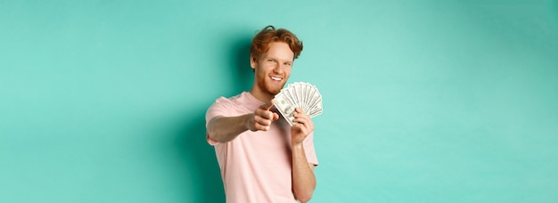 Foto gratuita chico pelirrojo guapo y confiado señalándote con el dedo mostrando dólares de dinero y concepto sonriente