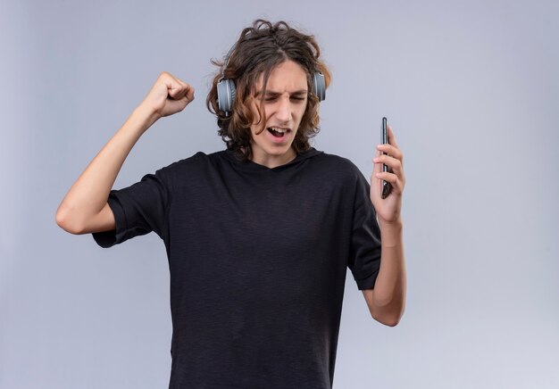 Chico con pelo largo en camiseta negra escucha música de auriculares en la pared blanca