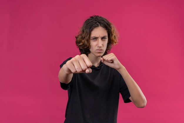 Chico con pelo largo en camiseta negra muestra el puño en la pared rosa