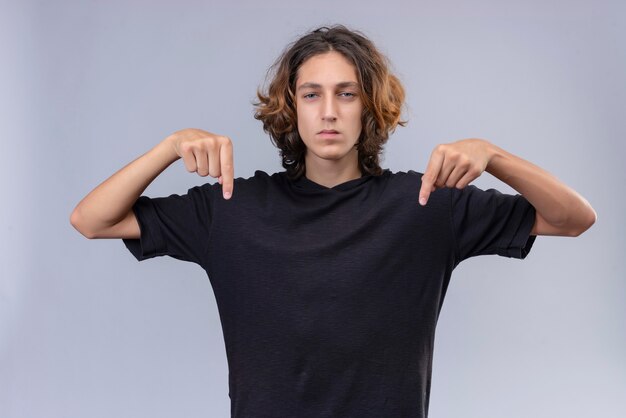 Chico con pelo largo en camiseta negra señala con los dedos hacia abajo en la pared blanca