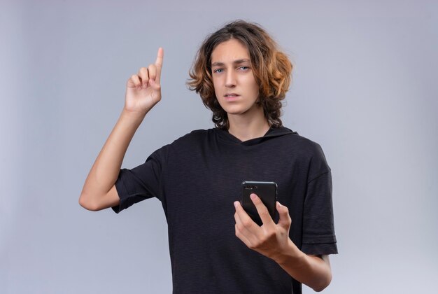 Chico con pelo largo en camiseta negra sosteniendo un teléfono y apunta hacia arriba en la pared blanca