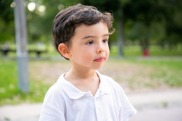 Chico de pelo negro lindo pensativo de pie en el parque de verano y mirando a otro lado. Fotografía de cerca. Concepto de infancia