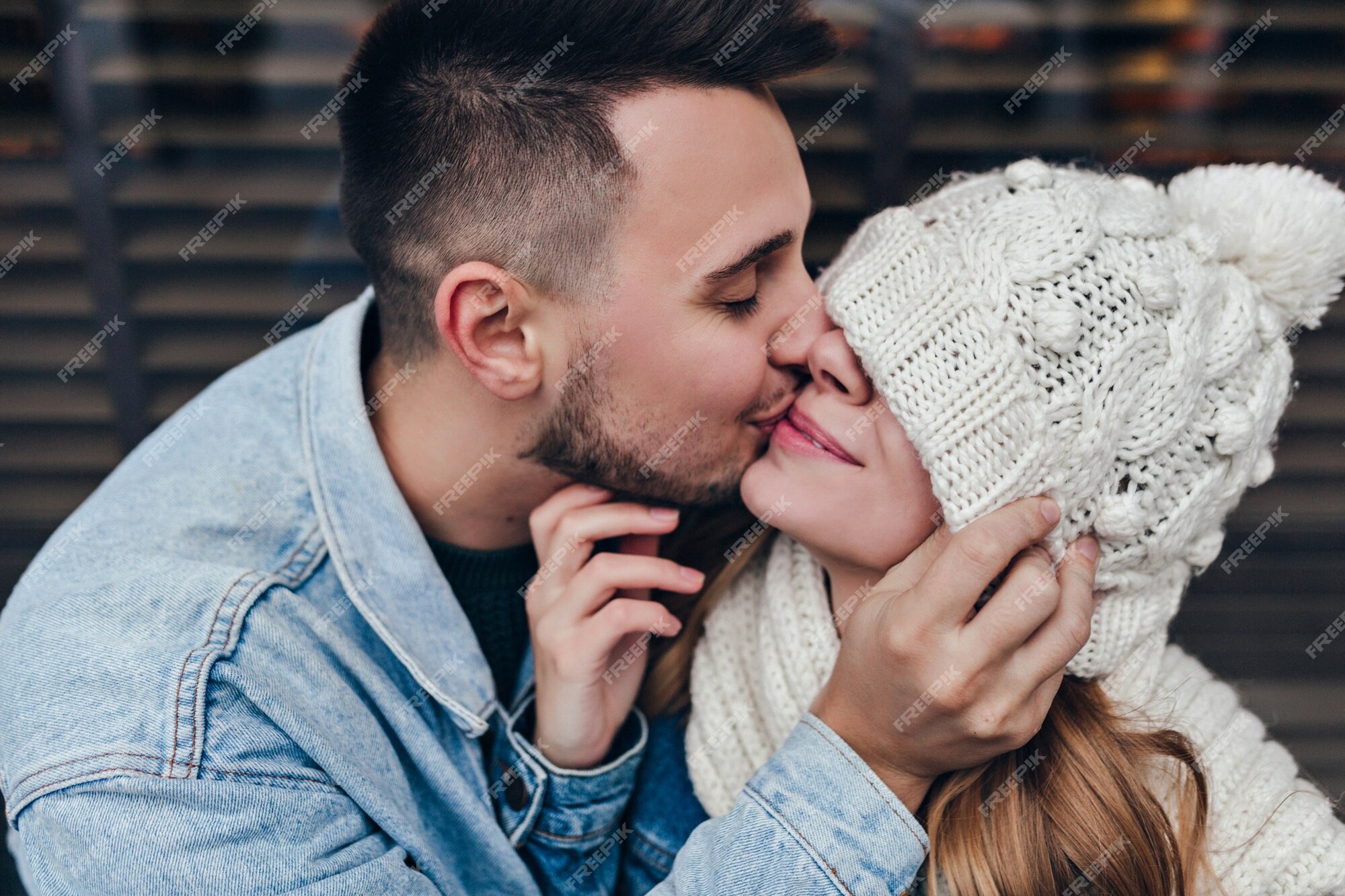 Chico de oscuro con chaqueta vaquera besando a su novia. hombre caucásico expresando a mujer divertida en gorro de invierno tejido. | Foto Gratis