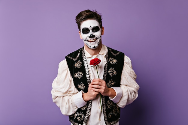 Chico de pelo oscuro con hermosa sonrisa de buen humor, posando en la pared aislada. foto de mexicano con arte facial y rosa en sus manos.