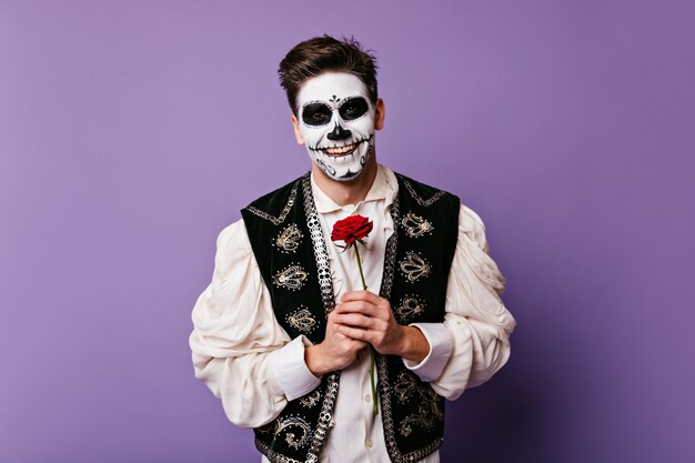 Chico de pelo oscuro con hermosa sonrisa de buen humor, posando en la pared aislada. Foto de mexicano con arte facial y rosa en sus manos.