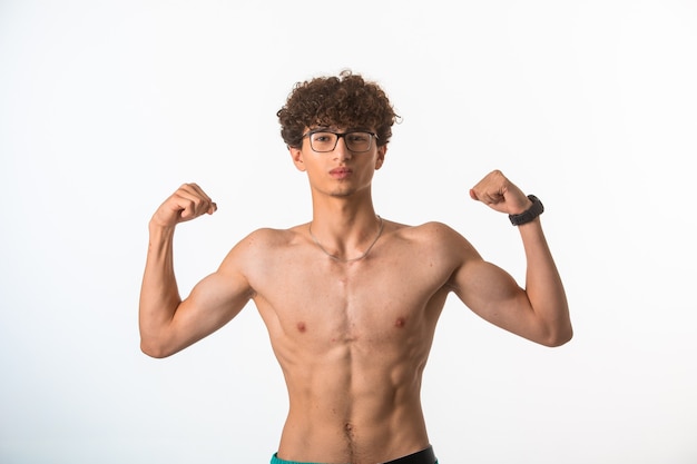 Chico de pelo rizado con gafas ópticas mostrando los músculos de su cuerpo en una posición desnuda.