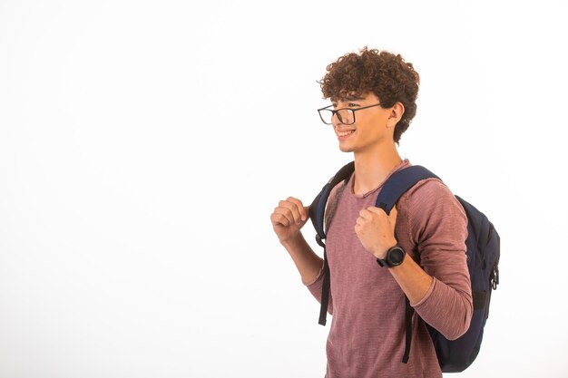 El chico de pelo rizado con gafas ópticas sosteniendo su mochila está seguro y motivado.