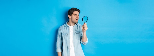 Foto gratuita chico pensativo joven mirando a través de una lupa leyendo algo pequeño de pie sobre la espalda azul