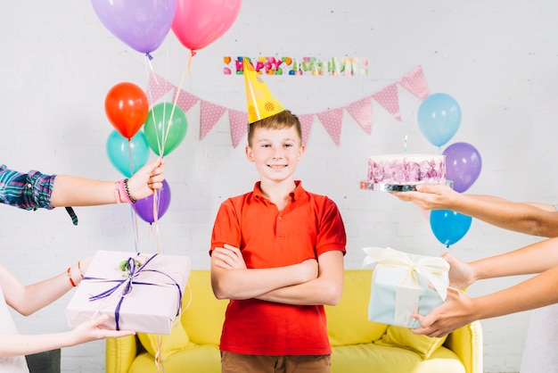 Chico de pie entre la mano de su amigo sosteniendo pastel de cumpleaños; regalos y globos