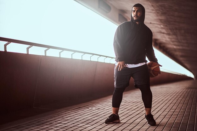 Chico de piel oscura vestido con una sudadera con capucha negra y pantalones cortos deportivos jugando baloncesto en una acera debajo de un puente.