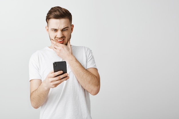 Chico preocupado encogerse ante la pantalla del teléfono inteligente, mirando ansioso al teléfono móvil