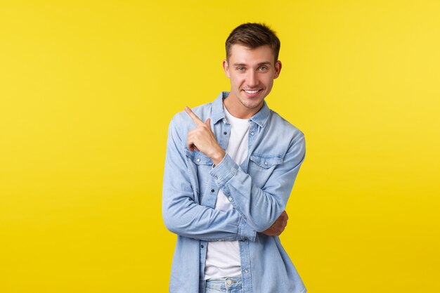 Chico queer guapo descarado y elegante con camisa de mezclilla, comprando para las vacaciones de verano, recogiendo ropa en la tienda, señalando la esquina superior izquierda, mostrando el enlace a la tienda en línea, fondo amarillo.