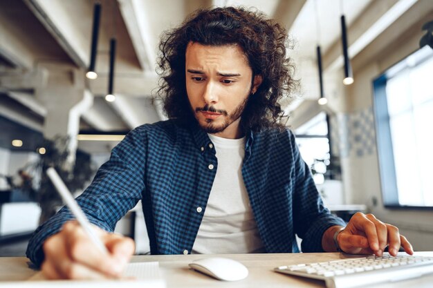 Chico rizado guapo está tomando notas en la mesa de trabajo en la oficina de espacios abiertos