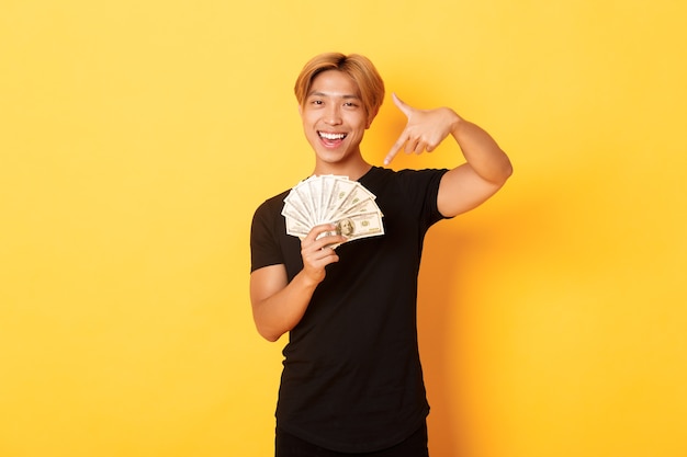 Chico rubio coreano guapo y descarado, sonriendo feliz y señalando con el dedo el dinero, ganando dinero en efectivo, pared amarilla de pie