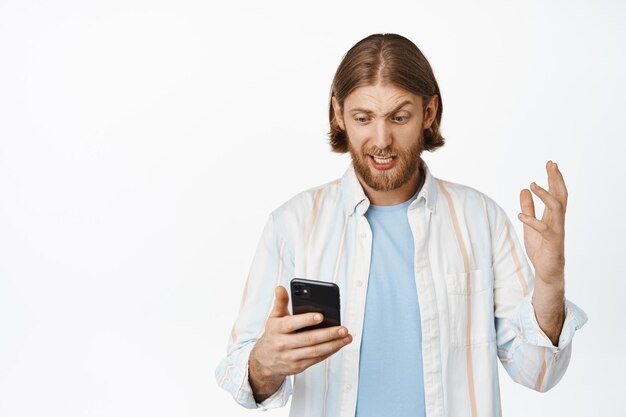 Chico rubio frustrado y confundido mirando el teléfono móvil con cara preocupada, leyendo malas noticias en el teléfono inteligente, de pie preocupado contra el fondo blanco