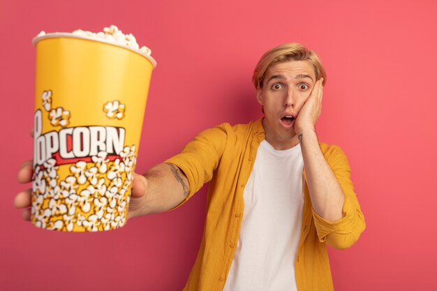 Chico rubio joven asustado con camiseta amarilla sosteniendo un cubo de palomitas de maíz