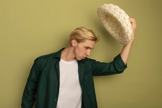 Chico rubio joven con camiseta verde poniendo sombrero en la cabeza.