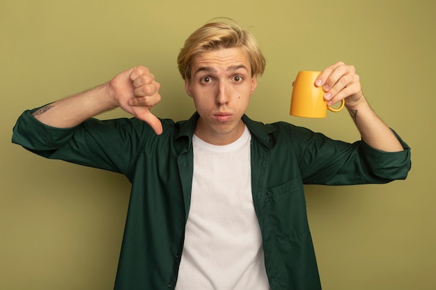 Chico rubio joven disgustado con camiseta verde sosteniendo la taza boca abajo mostrando el pulgar hacia abajo