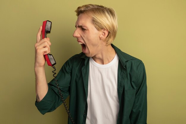 Chico rubio joven enojado con camiseta verde sosteniendo y mirando el teléfono