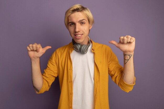 Chico rubio joven sonriente con camiseta amarilla y auriculares en el cuello apunta a sí mismo aislado en púrpura