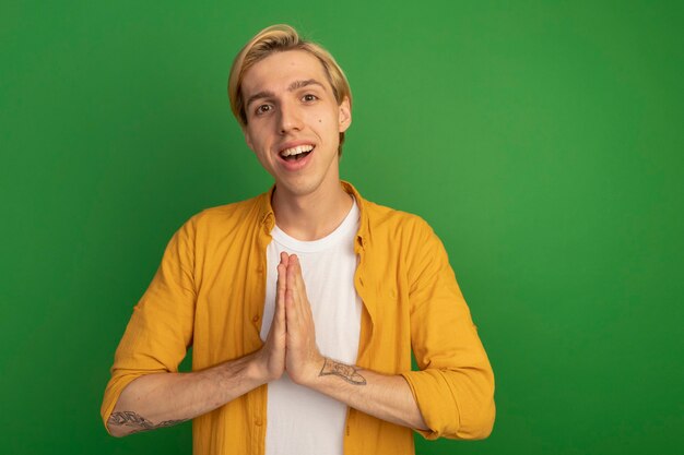 Chico rubio joven sonriente con camiseta amarilla que muestra gesto de oración aislado en verde con espacio de copia