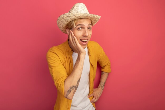 Foto gratuita chico rubio joven sonriente con camiseta amarilla y sombrero poniendo las manos en la mejilla y la cadera