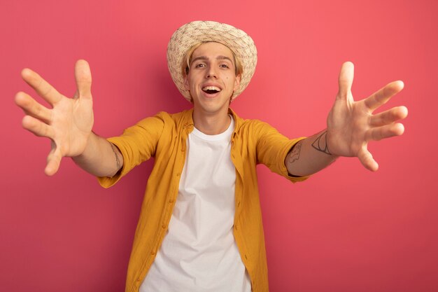 Chico rubio joven sonriente con camiseta amarilla y sombrero tomados de la mano aislados en rosa