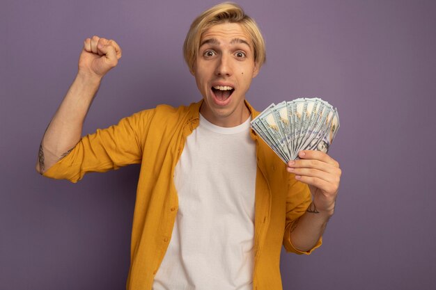 Chico rubio joven sonriente con camiseta amarilla sosteniendo dinero en efectivo y mostrando gesto sí aislado en púrpura