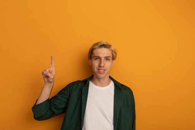 Chico rubio joven sonriente con camiseta verde apunta hacia arriba