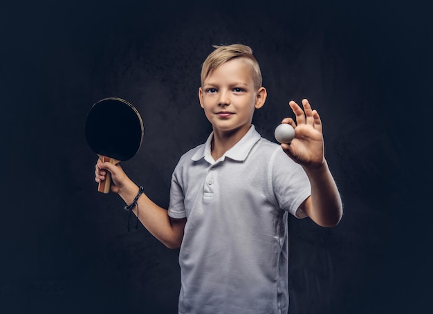 Un chico rubio vestido con una camiseta blanca sostiene una raqueta y una pelota de ping-pong en un estudio. Aislado en un fondo de textura oscura.