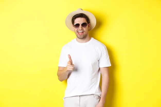 Chico seguro y descarado de vacaciones coqueteando contigo, señalando con el dedo a la cámara y guiñando un ojo, con sombrero de verano con gafas de sol, fondo amarillo