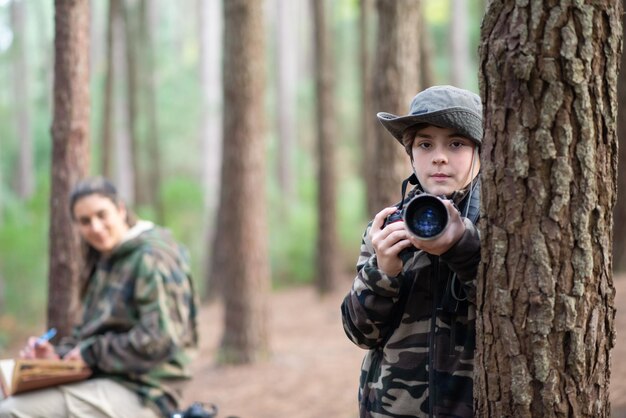 Chico serio con cámara en el bosque. Colegial con abrigo y panamá mirando a la cámara. madre borrosa en el fondo. Infancia, naturaleza, concepto de ocio.