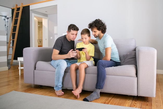 Chico serio jugando juegos en línea en el teléfono celular, sus dos papás sentados cerca de él y mirando la pantalla. Familia en casa y concepto de comunicación.